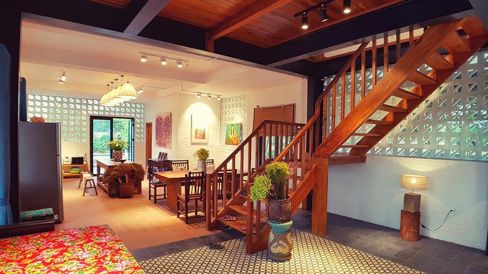 Khu bếp ăn nhìn từ phòng khách: cầu thang gỗ bậc hở, dầm bê tông sơn đen, tường gạch bông gió và bàn ăn gỗ dưới đèn mây; sàn zoning gạch bông – gạch đất nung