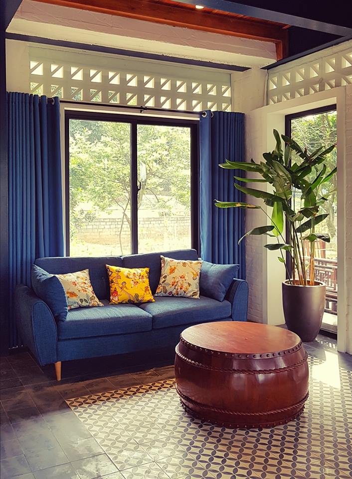 Góc sofa vải xanh navy tại Giang House, cửa kính lớn nhìn ra vườn, dải gạch bông gió cao sát trần, rèm xanh đậm, bàn trà tròn dáng trống gỗ và chậu cây nội thất trên nền “thảm” gạch bông.