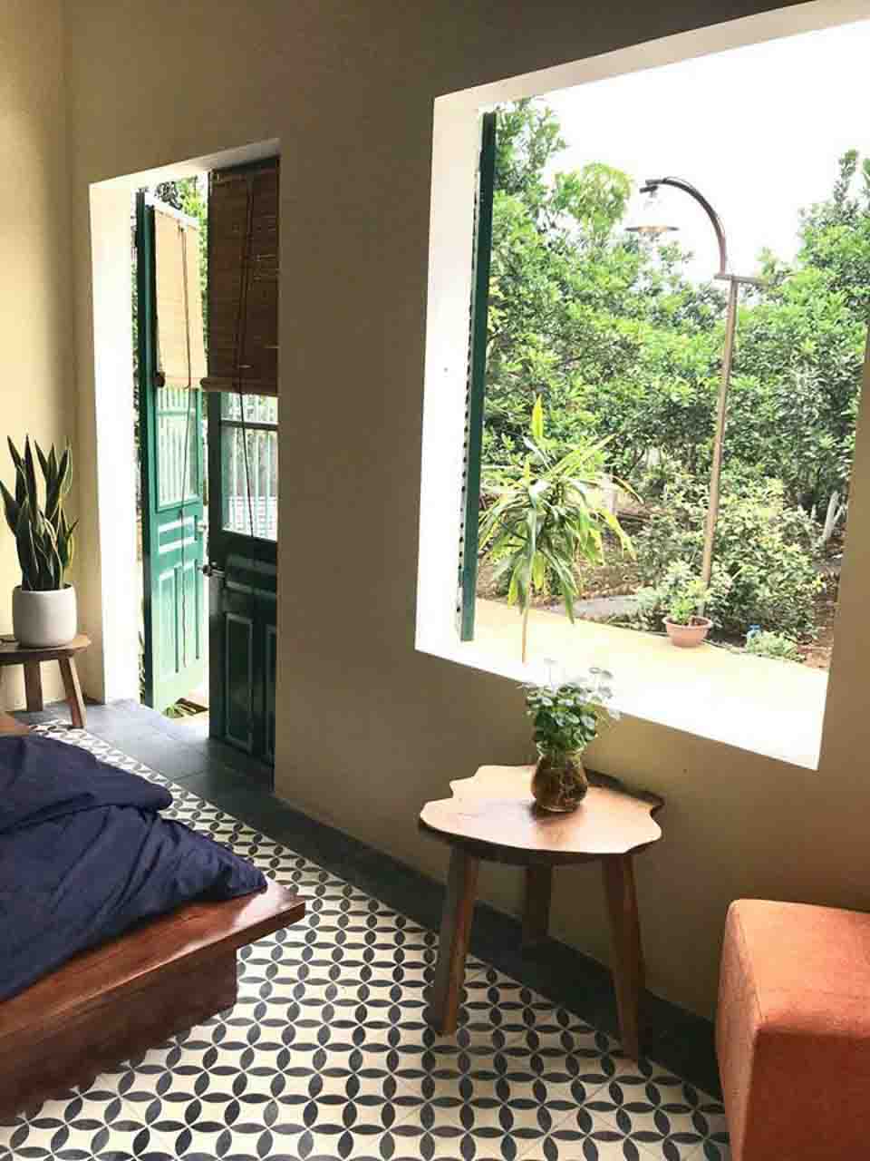 Góc phòng ngủ tầng 1 Giang House: nền gạch bông mát, bàn gỗ mộc đặt ấm trà, giường gỗ platform, cửa mở ra vườn và ô cửa sổ lớn lấy sáng tự nhiên.