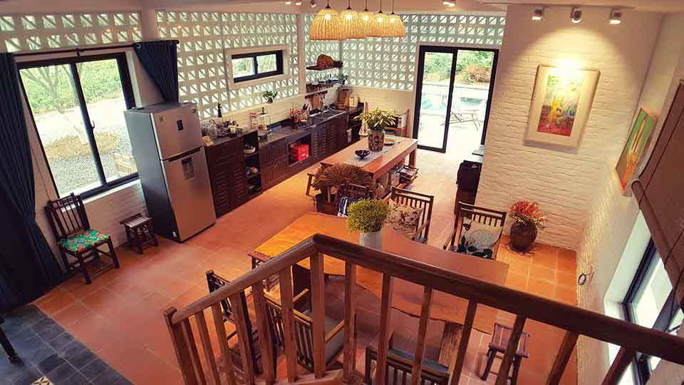 Tổng thể bếp ăn với dãy tủ bếp áp tường, bàn soạn ở giữa, đèn mây thả, cửa kính mở ra hiên hồ bơi; tường gạch bông gió và sàn gạch đất nung.