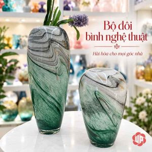 Hai dáng bình thủy tinh vân đá xanh ngọc đặt cạnh nhau, bình cao cắm hoa tím