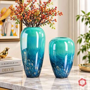 Hai bình thủy tinh chuyển sắc xanh đặt cạnh nhau trên bàn đá sáng, bình cao cắm cành quả đỏ cam