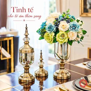 Bình thủy tinh cắm hoa xanh trắng, đặt bên hai vật trang trí ánh kim trên bàn ăn