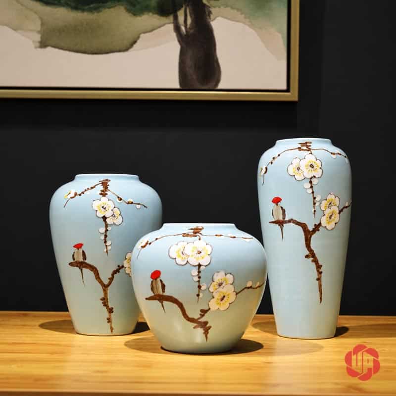 Bộ Bình Gốm Sứ Hoa Mai Trắng 3 Món Trang Trí Phòng Khách BG023 Bộ ba bình gốm hoa mai trắng xanh ngọc đặt trên bàn gỗ dưới tranh treo tường nền tối.