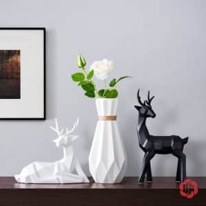 Tượng hươu trắng nằm cạnh bình hoa đa giác và tượng hươu đen đứng trên tủ gỗ.